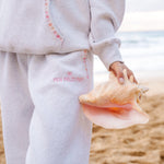 Matching heather grey sweat set with Pink Palm Puff shell pattern border. Walking toward the beach, shot from behind. Overcast soft light, cool tones, wet sand reflection, muted ocean horizon. Aesthetic beach editorial, film grain.
