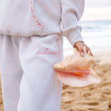 Matching heather grey sweat set with Pink Palm Puff shell pattern border. Walking toward the beach, shot from behind. Overcast soft light, cool tones, wet sand reflection, muted ocean horizon. Aesthetic beach editorial, film grain.