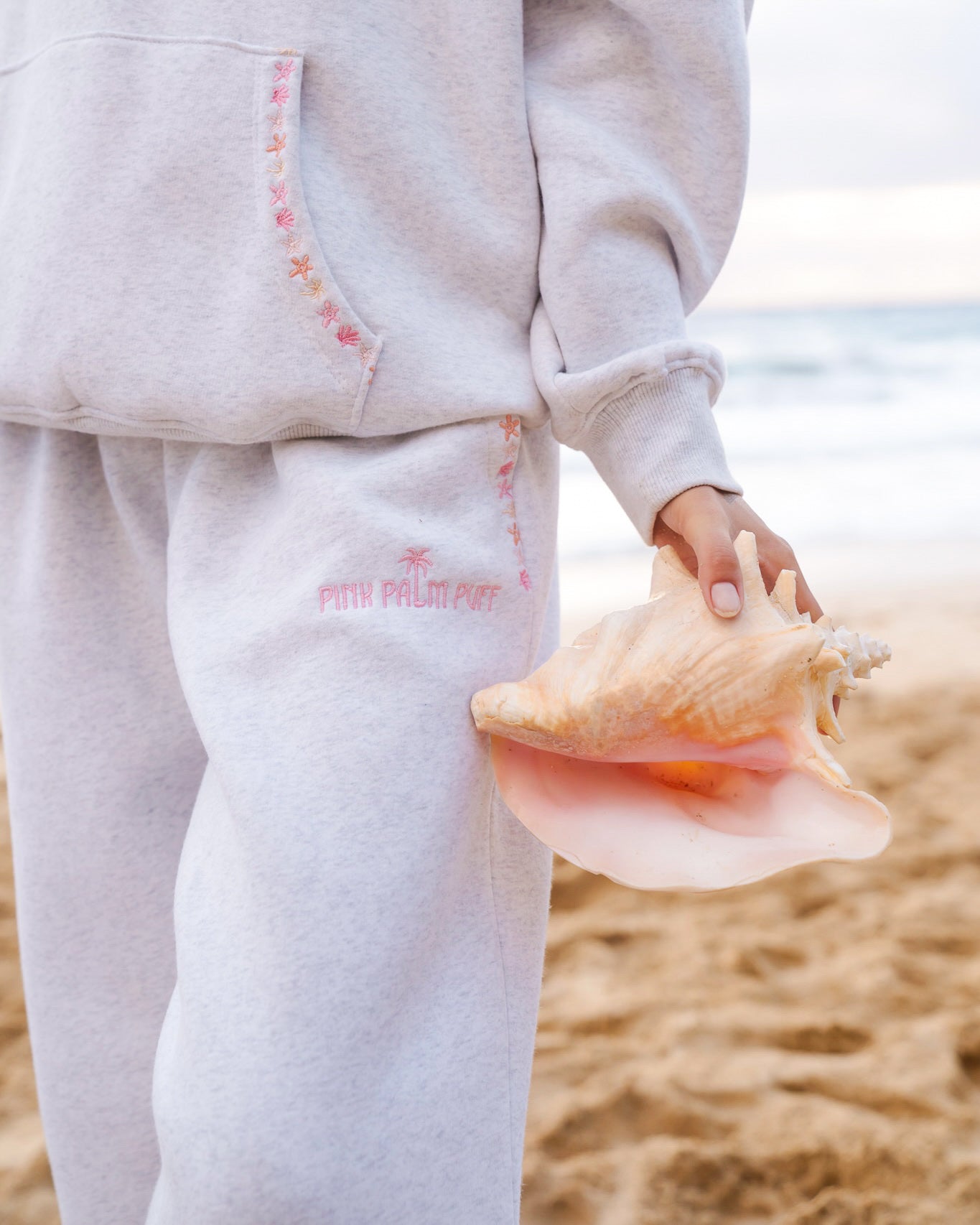 Matching heather grey sweat set with Pink Palm Puff shell pattern border. Walking toward the beach, shot from behind. Overcast soft light, cool tones, wet sand reflection, muted ocean horizon. Aesthetic beach editorial, film grain.