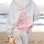Matching heather grey sweat set with Pink Palm Puff shell pattern border. Walking toward the beach, shot from behind. Overcast soft light, cool tones, wet sand reflection, muted ocean horizon. Aesthetic beach editorial, film grain.