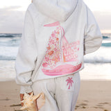 Matching heather grey sweat set with Pink Palm Puff shell pattern border. Walking toward the beach, shot from behind. Overcast soft light, cool tones, wet sand reflection, muted ocean horizon. Aesthetic beach editorial, film grain.