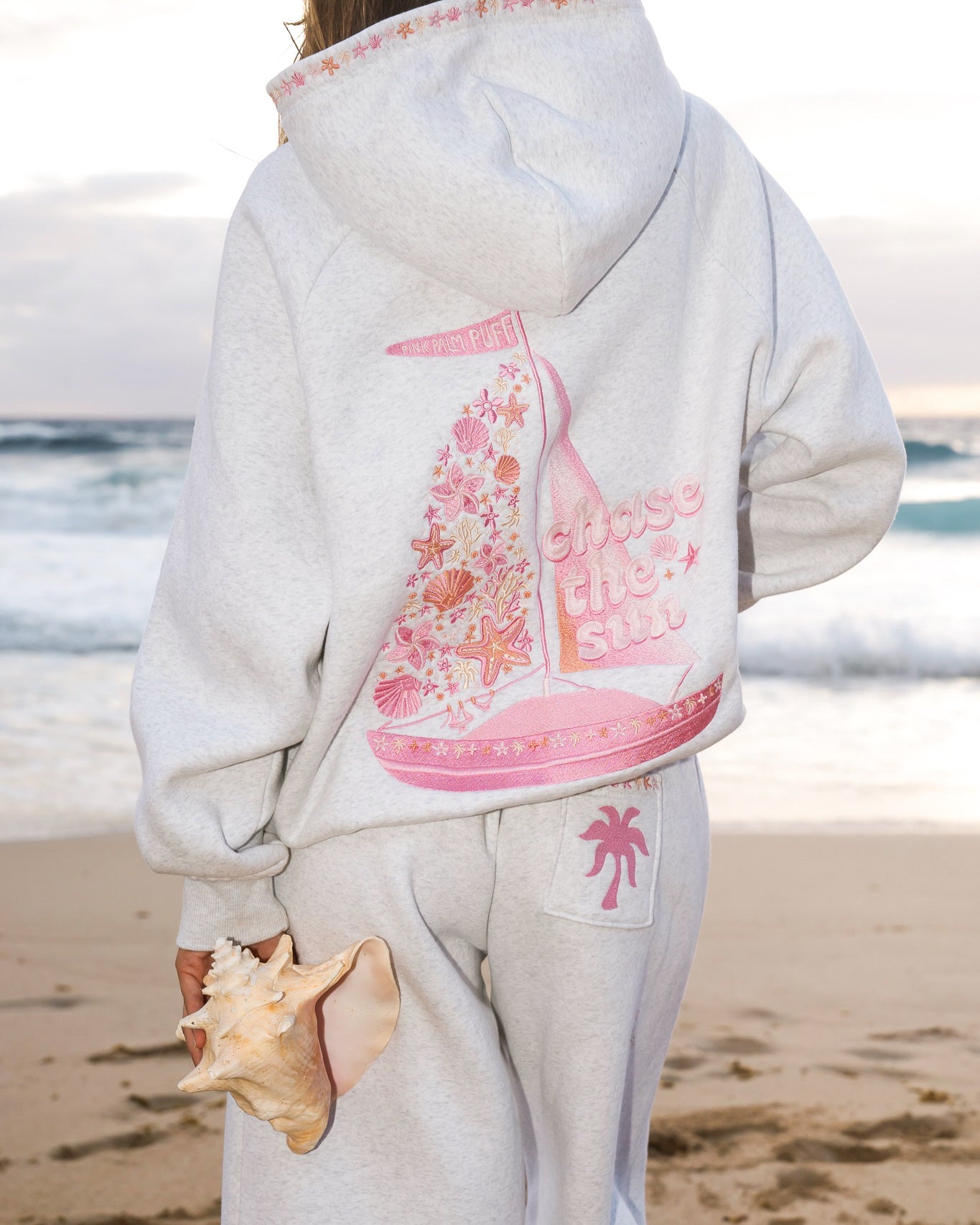 Matching heather grey sweat set with Pink Palm Puff shell pattern border. Walking toward the beach, shot from behind. Overcast soft light, cool tones, wet sand reflection, muted ocean horizon. Aesthetic beach editorial, film grain.