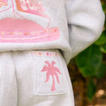 Matching heather grey sweat set with Pink Palm Puff shell pattern border. Walking toward the beach, shot from behind. Overcast soft light, cool tones, wet sand reflection, muted ocean horizon. Aesthetic beach editorial, film grain.