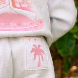 Matching heather grey sweat set with Pink Palm Puff shell pattern border. Walking toward the beach, shot from behind. Overcast soft light, cool tones, wet sand reflection, muted ocean horizon. Aesthetic beach editorial, film grain.