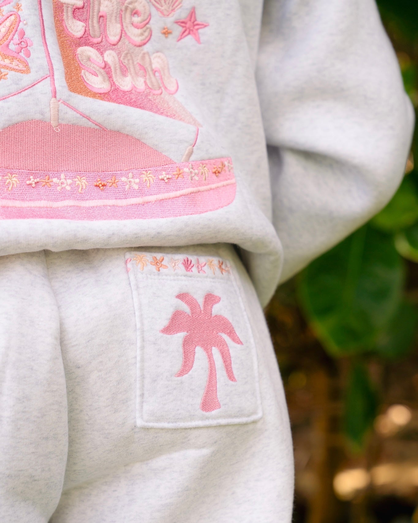 Matching heather grey sweat set with Pink Palm Puff shell pattern border. Walking toward the beach, shot from behind. Overcast soft light, cool tones, wet sand reflection, muted ocean horizon. Aesthetic beach editorial, film grain.