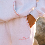 Matching heather grey sweat set with Pink Palm Puff shell pattern border. Walking toward the beach, shot from behind. Overcast soft light, cool tones, wet sand reflection, muted ocean horizon. Aesthetic beach editorial, film grain.