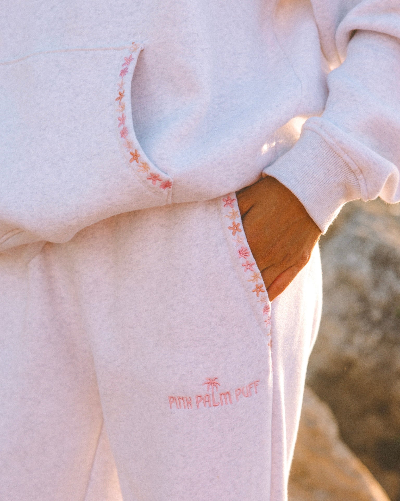 Matching heather grey sweat set with Pink Palm Puff shell pattern border. Walking toward the beach, shot from behind. Overcast soft light, cool tones, wet sand reflection, muted ocean horizon. Aesthetic beach editorial, film grain.