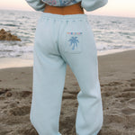 Matching powder blue sweat set with Pink Palm Puff shell pattern border. GWalking toward the beach, shot from behind. Golden hour, soft sand, ocean and low dunes in frame. Aesthetic beach editorial, warm natural light, film grain.