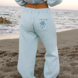 Matching powder blue sweat set with Pink Palm Puff shell pattern border. GWalking toward the beach, shot from behind. Golden hour, soft sand, ocean and low dunes in frame. Aesthetic beach editorial, warm natural light, film grain.
