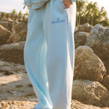Matching powder blue sweat set with Pink Palm Puff shell pattern border. GWalking toward the beach, shot from behind. Golden hour, soft sand, ocean and low dunes in frame. Aesthetic beach editorial, warm natural light, film grain.