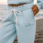Matching powder blue sweat set with Pink Palm Puff shell pattern border. GWalking toward the beach, shot from behind. Golden hour, soft sand, ocean and low dunes in frame. Aesthetic beach editorial, warm natural light, film grain.