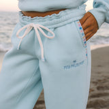Matching powder blue sweat set with Pink Palm Puff shell pattern border. GWalking toward the beach, shot from behind. Golden hour, soft sand, ocean and low dunes in frame. Aesthetic beach editorial, warm natural light, film grain.