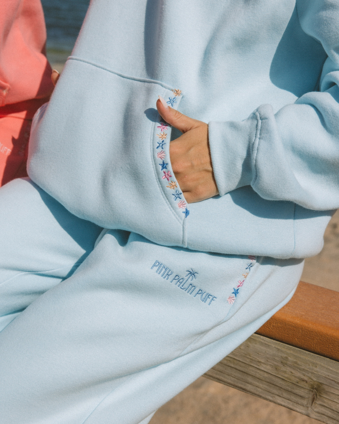 Matching powder blue sweat set with Pink Palm Puff shell pattern border. GWalking toward the beach, shot from behind. Golden hour, soft sand, ocean and low dunes in frame. Aesthetic beach editorial, warm natural light, film grain.