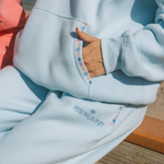 Matching powder blue sweat set with Pink Palm Puff shell pattern border. GWalking toward the beach, shot from behind. Golden hour, soft sand, ocean and low dunes in frame. Aesthetic beach editorial, warm natural light, film grain.