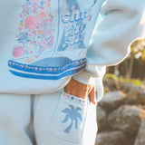 Matching powder blue sweat set with Pink Palm Puff shell pattern border. GWalking toward the beach, shot from behind. Golden hour, soft sand, ocean and low dunes in frame. Aesthetic beach editorial, warm natural light, film grain.