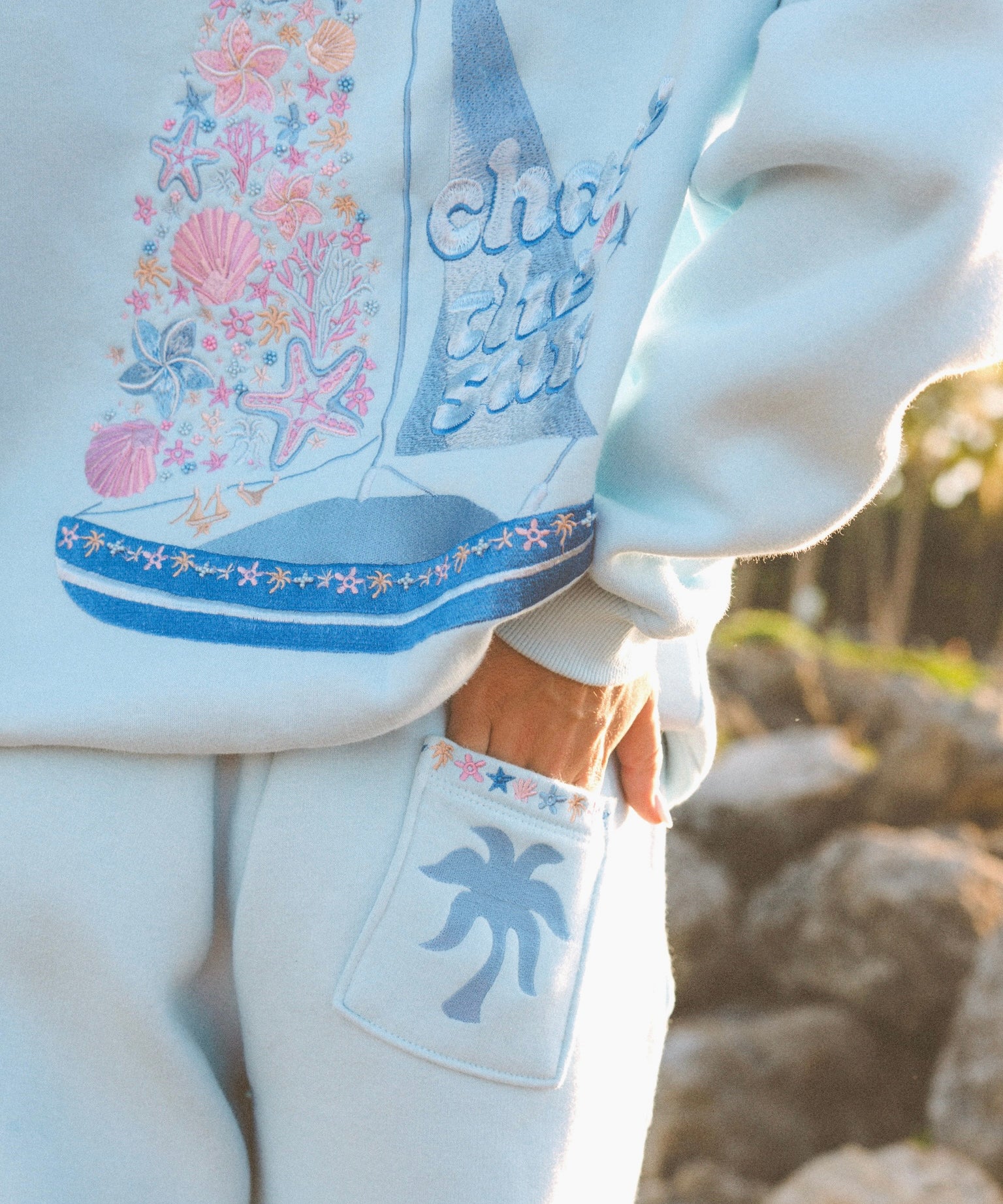 Matching powder blue sweat set with Pink Palm Puff shell pattern border. GWalking toward the beach, shot from behind. Golden hour, soft sand, ocean and low dunes in frame. Aesthetic beach editorial, warm natural light, film grain.