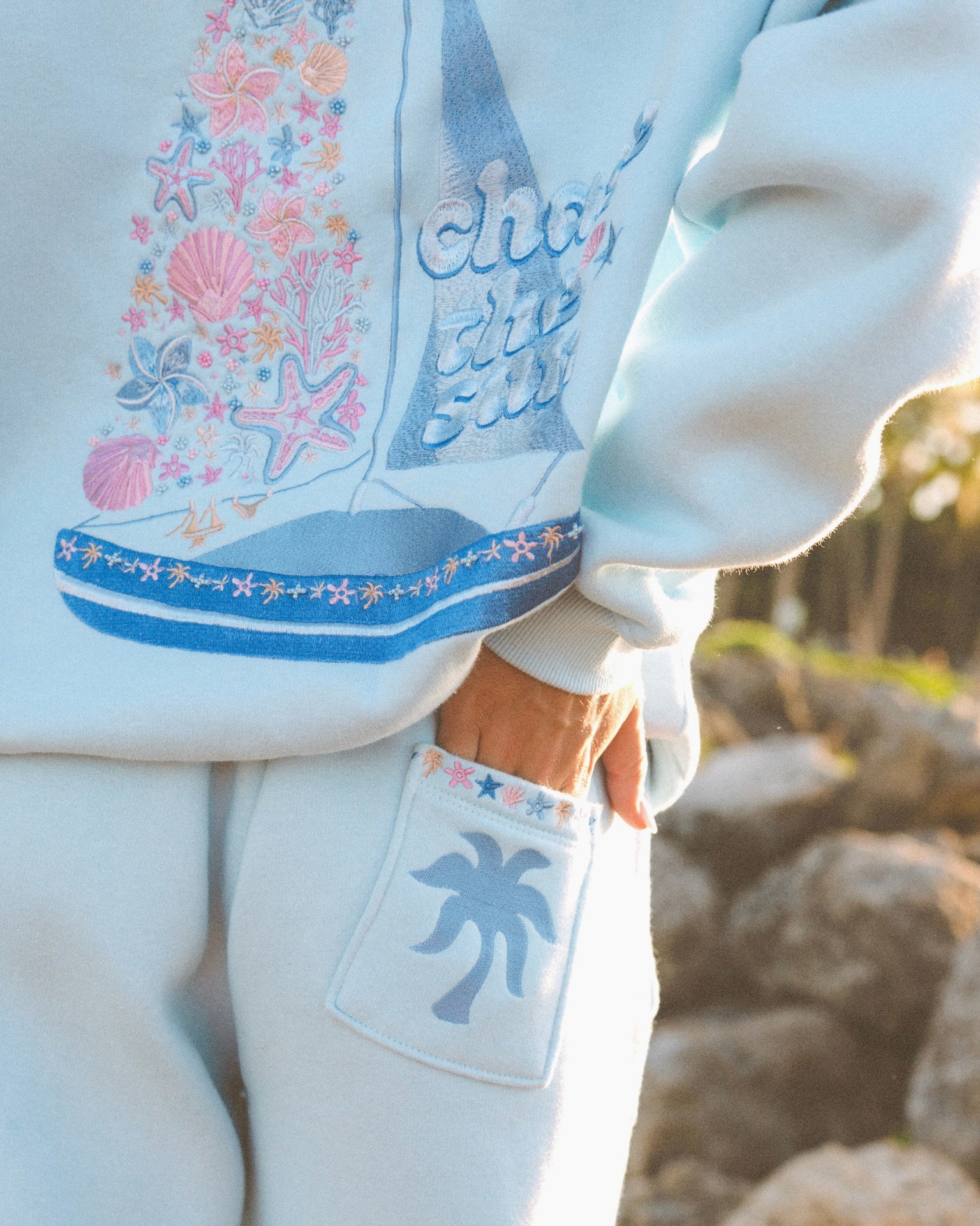 Matching powder blue sweat set with Pink Palm Puff shell pattern border. GWalking toward the beach, shot from behind. Golden hour, soft sand, ocean and low dunes in frame. Aesthetic beach editorial, warm natural light, film grain.