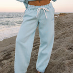 Matching powder blue sweat set with Pink Palm Puff shell pattern border. GWalking toward the beach, shot from behind. Golden hour, soft sand, ocean and low dunes in frame. Aesthetic beach editorial, warm natural light, film grain.