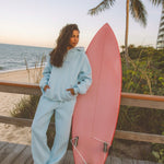 Matching powder blue sweat set with Pink Palm Puff shell pattern border. GWalking toward the beach, shot from behind. Golden hour, soft sand, ocean and low dunes in frame. Aesthetic beach editorial, warm natural light, film grain.