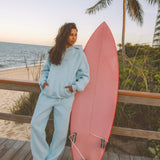 Matching powder blue sweat set with Pink Palm Puff shell pattern border. GWalking toward the beach, shot from behind. Golden hour, soft sand, ocean and low dunes in frame. Aesthetic beach editorial, warm natural light, film grain.