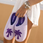 Person holding a pair of purple Malibu slippers by Pink Palm Puff with palm tree design on a blurred background