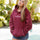 Woman wearing a maroon Pink Palm Puff hoodie with text, standing outdoors with greenery in the background